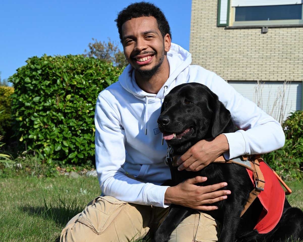 Un jeune homme souriant, assis dans un jardin, tient contre lui un chien guide noir portant un harnais.