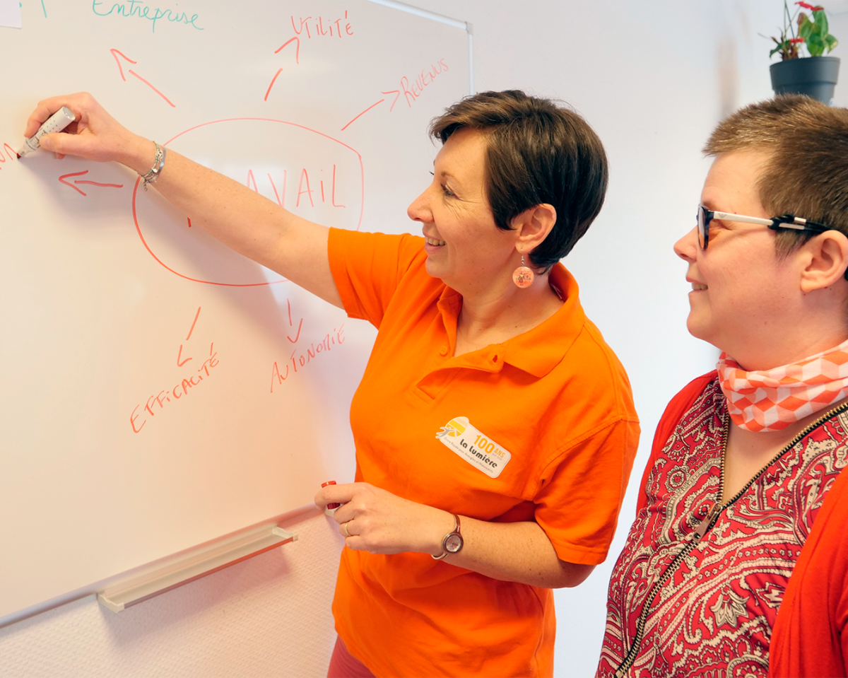 Deux femmes échangent devant un tableau blanc. L'une d'elles, vêtue d'un polo orange, présente un schéma.