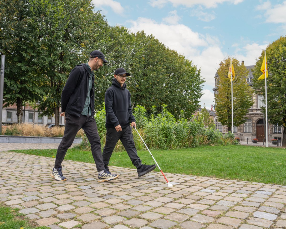 Deux hommes marchent sur un chemin pavé dans un parc. L'un d'eux utilise une canne blanche pour se déplacer.